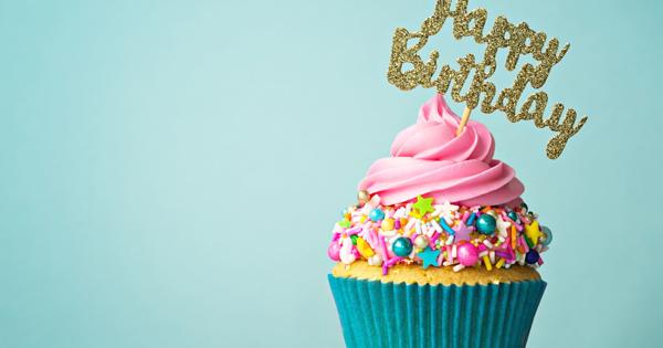Cupcake with pink frosting, colorful sprinkles, and candies in a blue wrapper, topped with a gold glittery 'Happy Birthday' sign. Set against a light blue background with sprinkles scattered around.