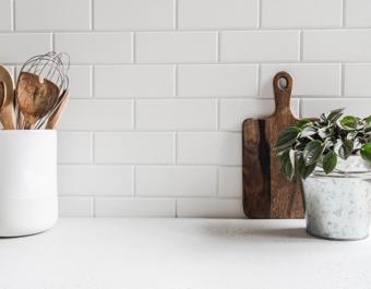 Minimalist kitchen countertop with white subway tile backsplash, wooden utensils in a ceramic container, a cutting board, and a small potted plant.