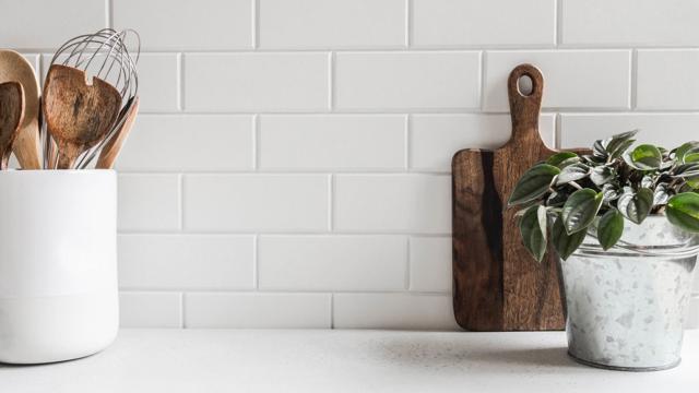 Minimalist kitchen countertop with white subway tile backsplash, wooden utensils in a ceramic container, a cutting board, and a small potted plant.