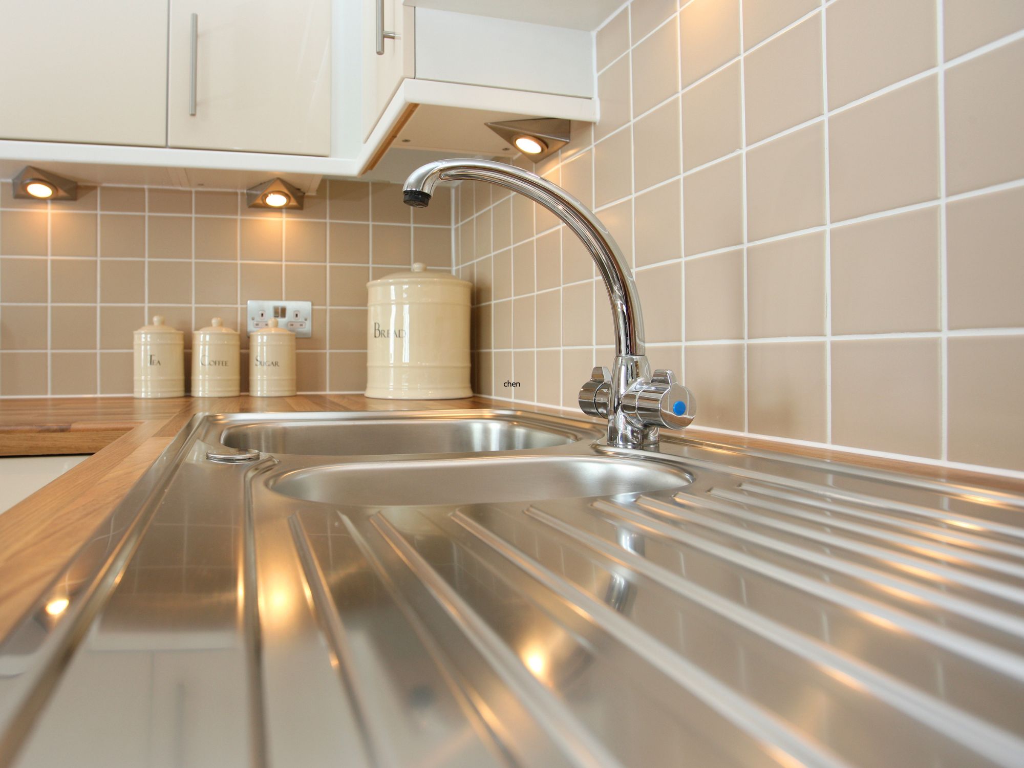 Modern kitchen sink area with a stainless steel sink, wooden countertop, beige tiled backsplash, and ceramic canisters labeled 'Tea,' 'Coffee,' 'Sugar,' and 'Biscuits' under cabinet spotlights.