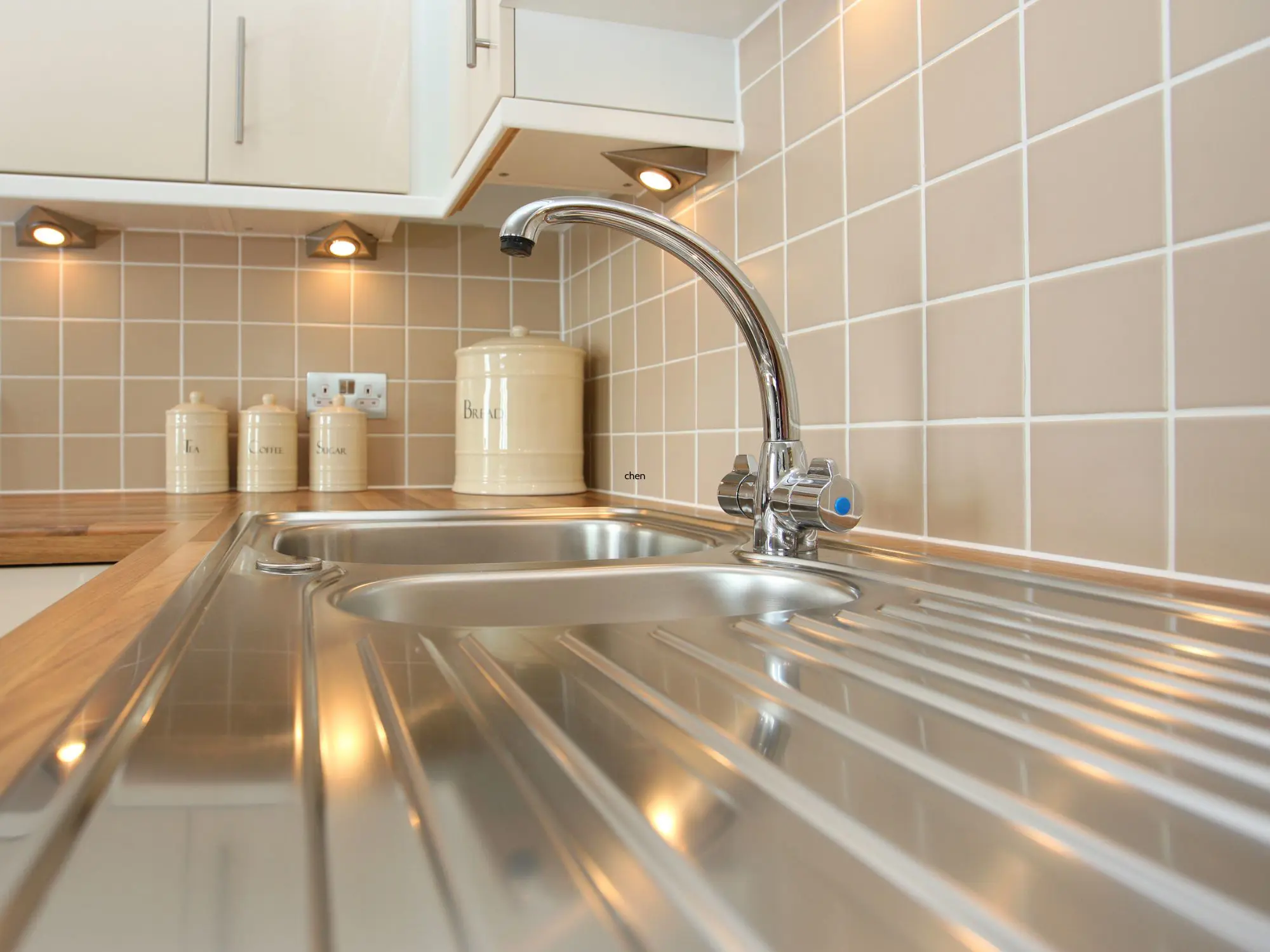 Modern kitchen sink area with a stainless steel sink, wooden countertop, beige tiled backsplash, and ceramic canisters labeled 'Tea,' 'Coffee,' 'Sugar,' and 'Biscuits' under cabinet spotlights.