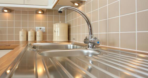 Modern kitchen sink area with a stainless steel sink, wooden countertop, beige tiled backsplash, and ceramic canisters labeled 'Tea,' 'Coffee,' 'Sugar,' and 'Biscuits' under cabinet spotlights.