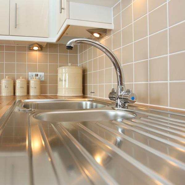 Modern kitchen sink area with a stainless steel sink, wooden countertop, beige tiled backsplash, and ceramic canisters labeled 'Tea,' 'Coffee,' 'Sugar,' and 'Biscuits' under cabinet spotlights.